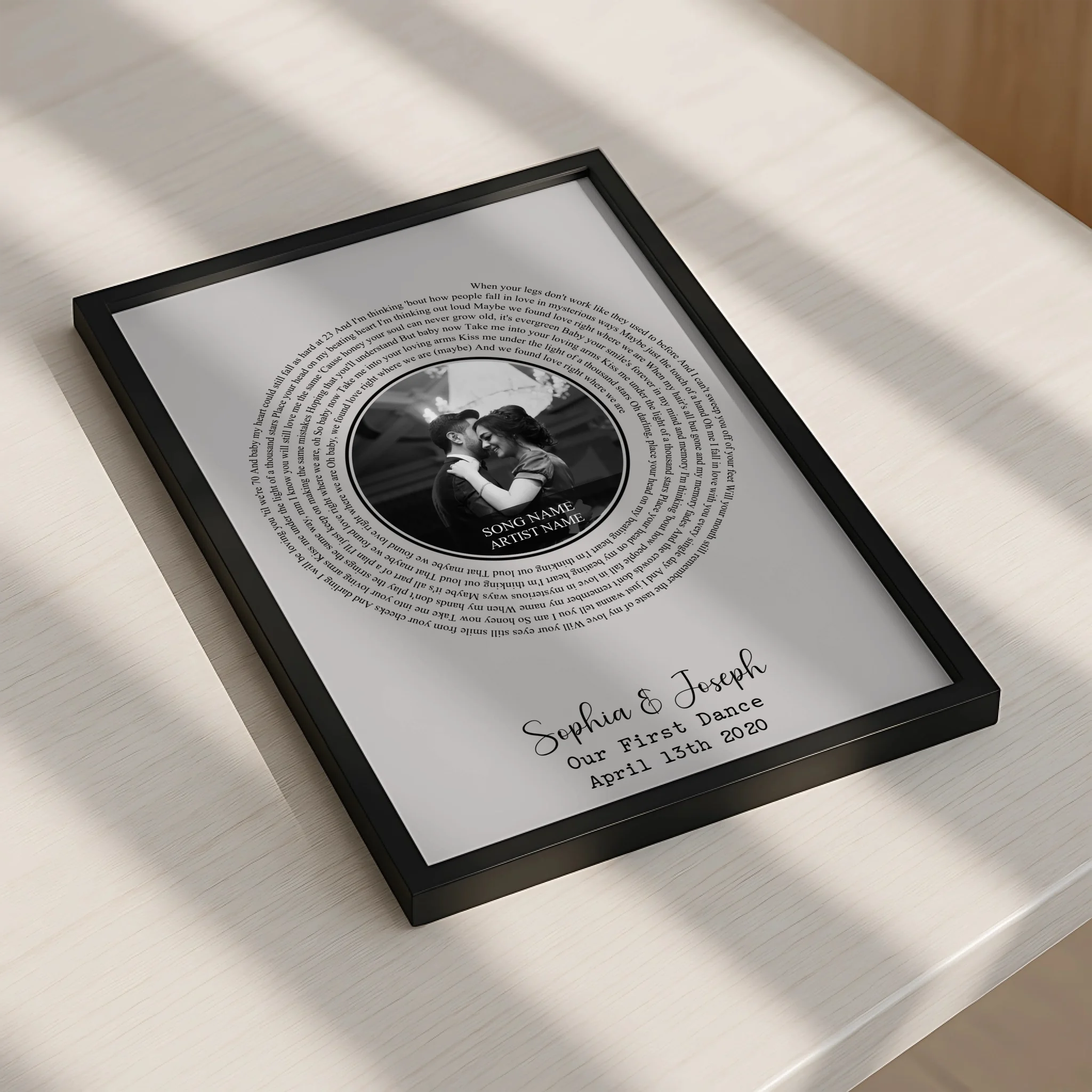 Black framed spiral song lyric print laid flat on a white surface in soft window light, showing couple's photo and Sophia and