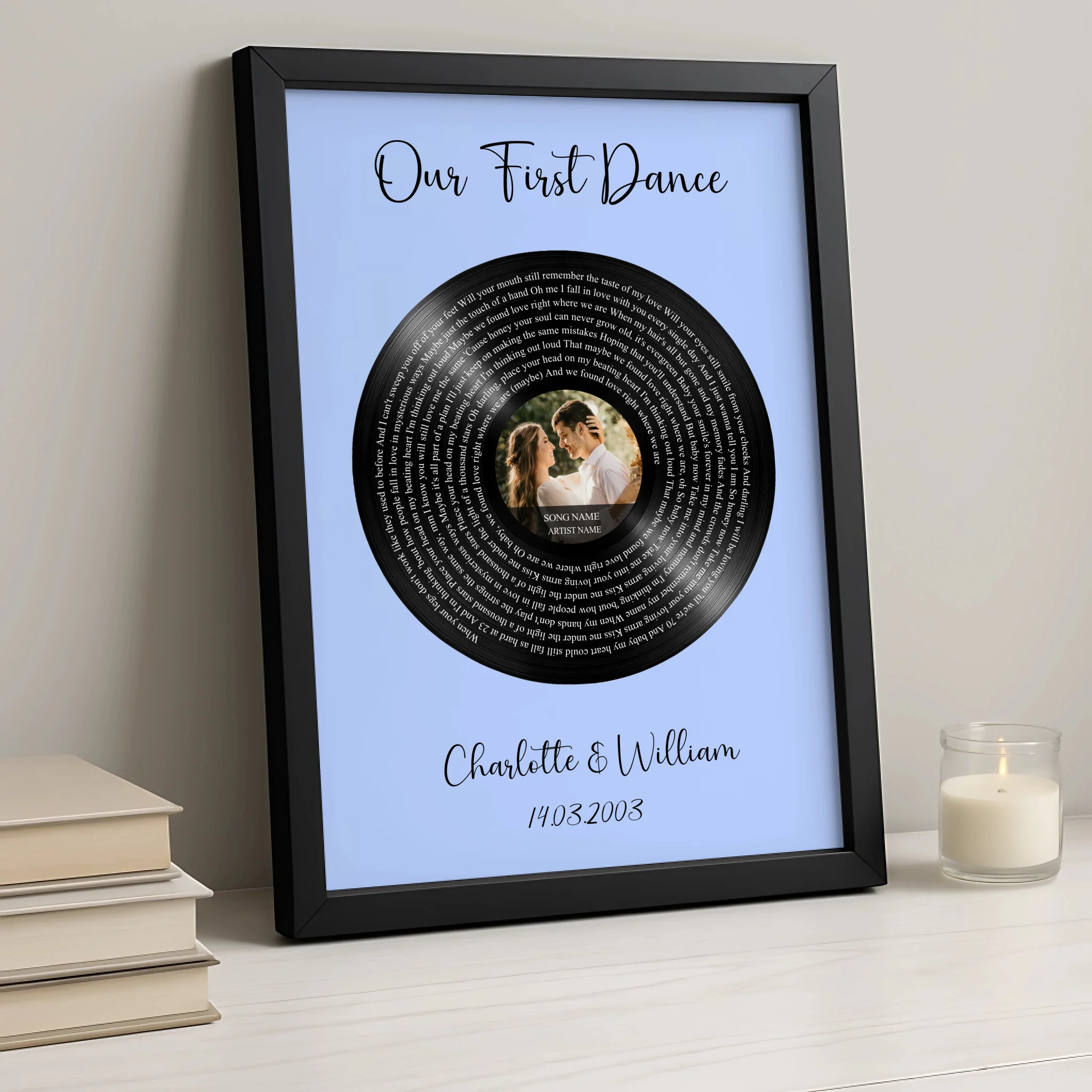 Black framed Our First Dance lyrics print on blue background, vinyl record with couple photo, beside stacked books and lit ca