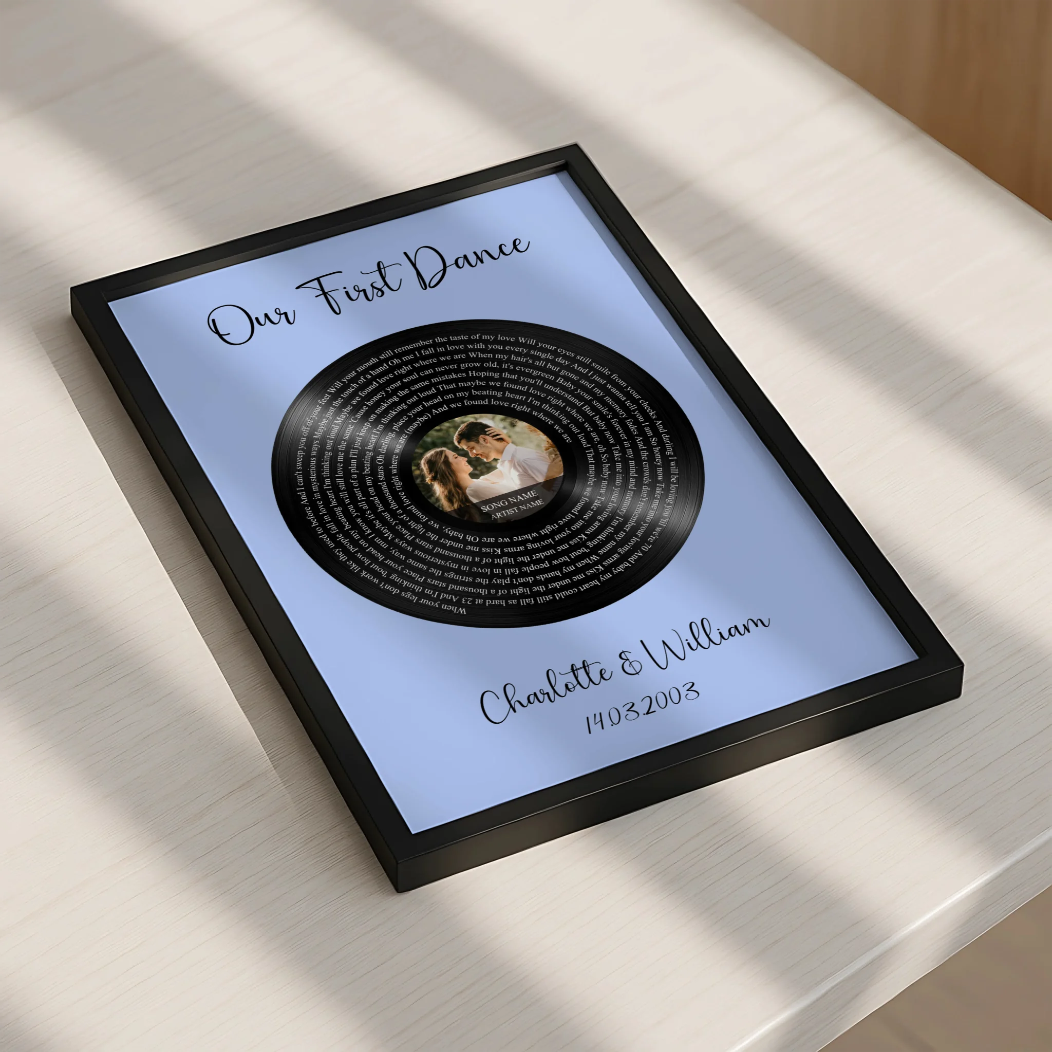 Black framed personalised song lyrics vinyl print on blue, lying flat on a sunlit white surface, showing Charlotte and Willia