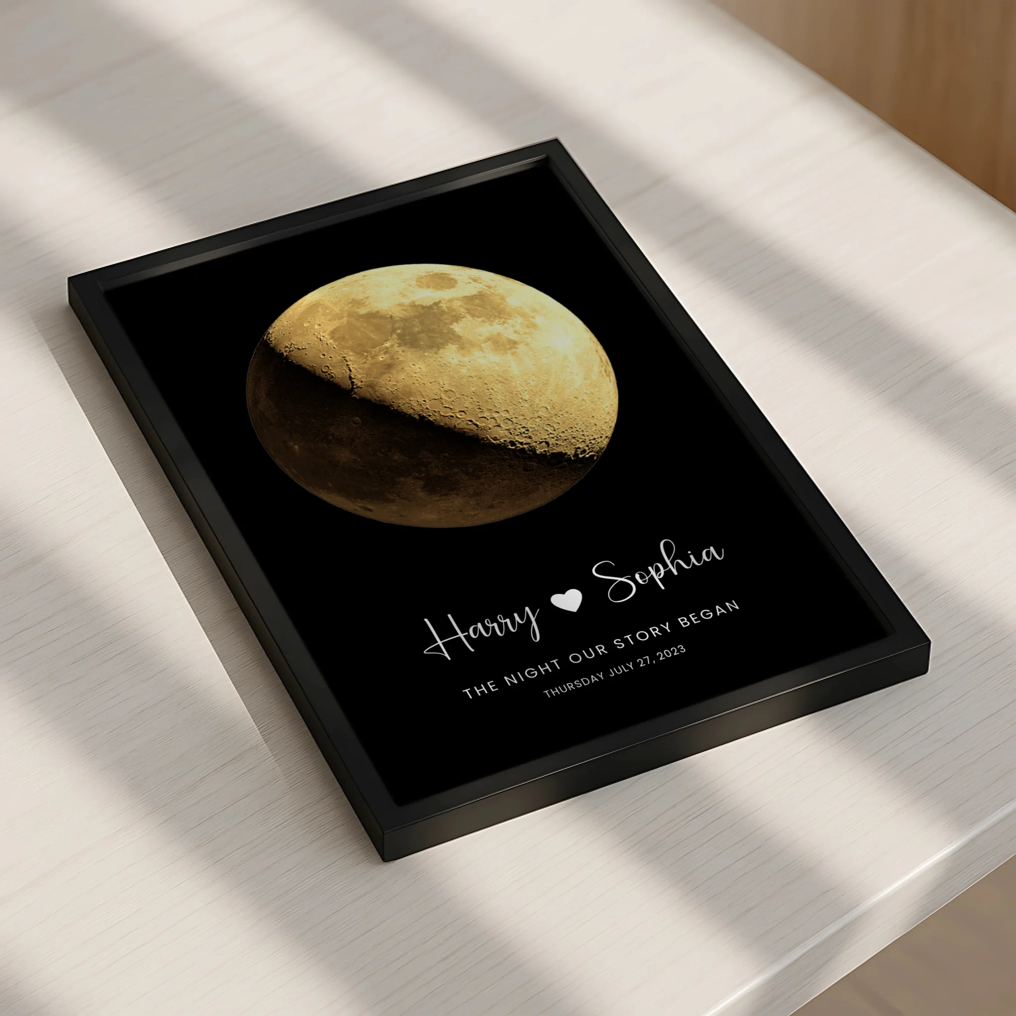 Personalised moon phase print in black frame lying flat on a table, showing gold waning gibbous moon, names Harry and Sophia,