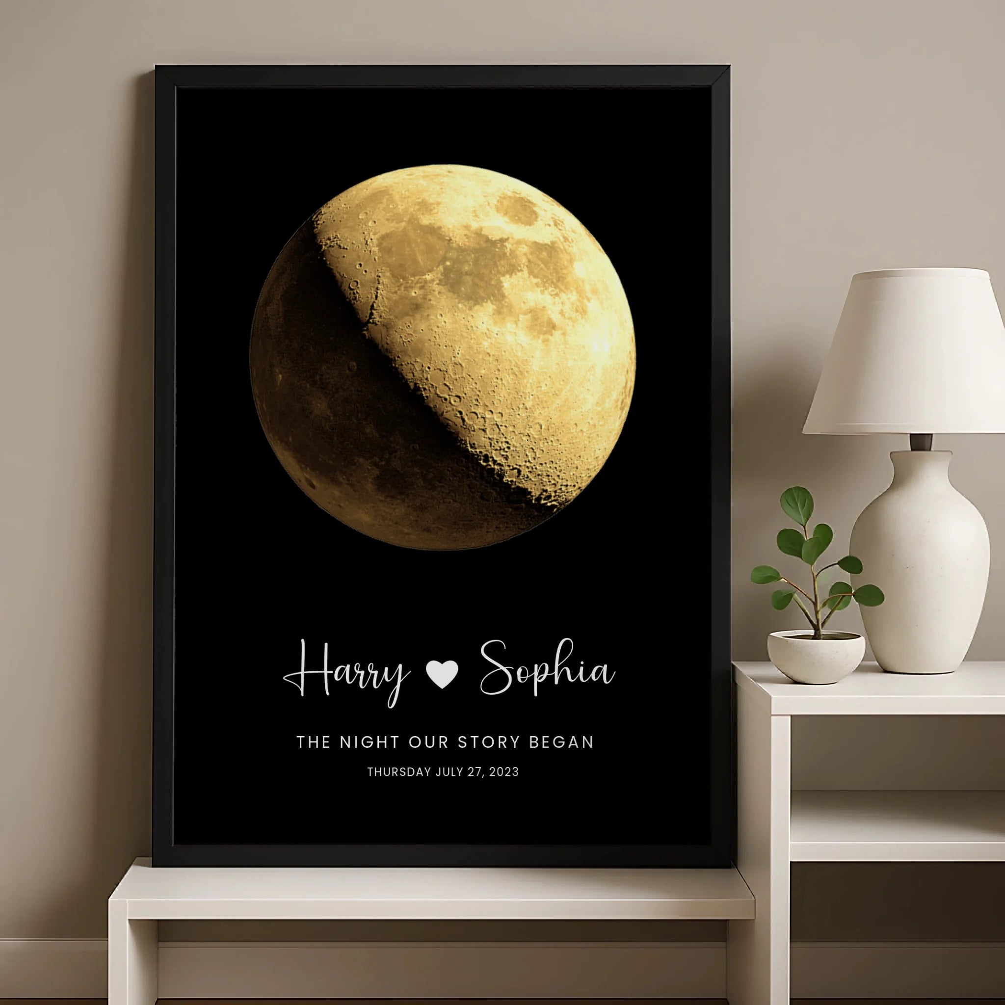 Black framed moon phase print for Harry and Sophia dated July 27 2023, leaning against a wall on a white sideboard next to a