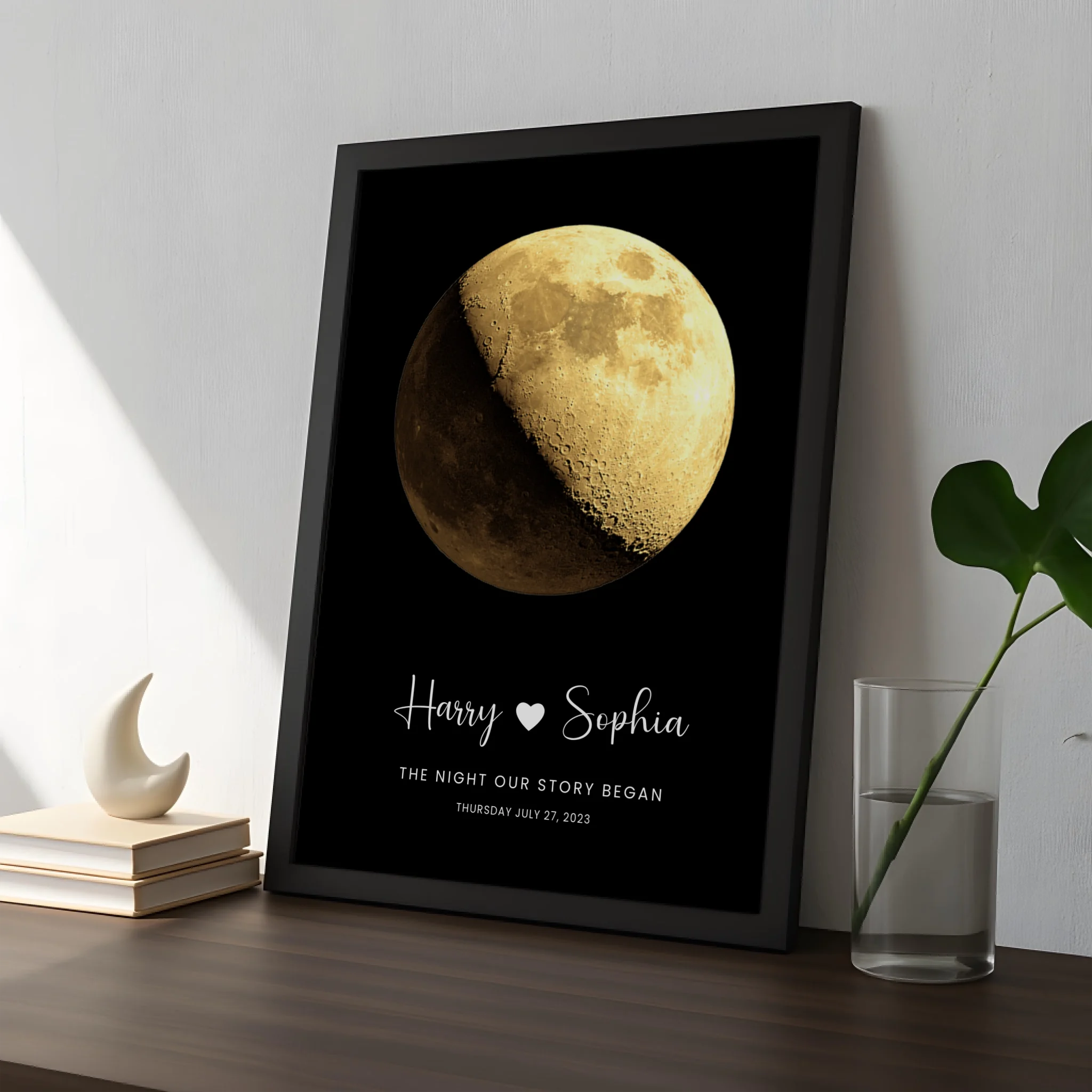 Black framed moon phase print personalised for Harry and Sophia, July 27 2023, propped on a dark wooden shelf beside a cresce
