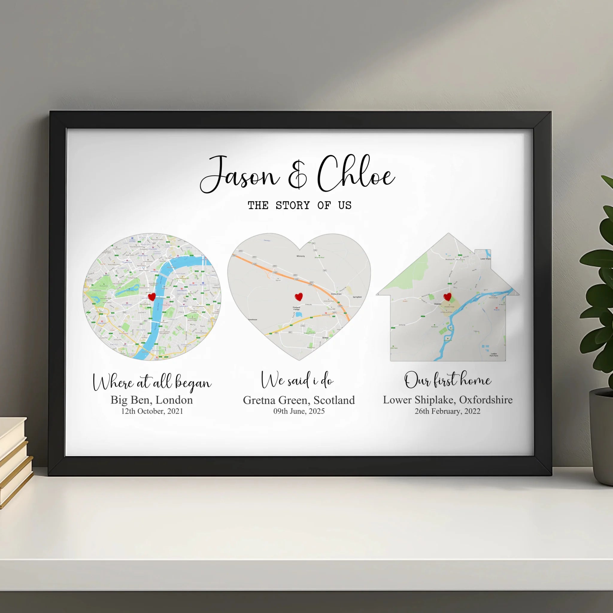 Black framed Story of Us couples map print on a white shelf beside stacked books and a small plant, three shaped map panels.