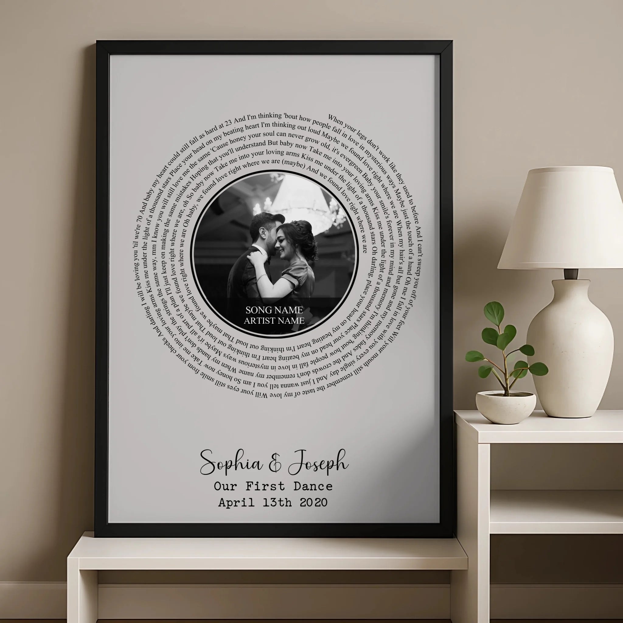 Large black framed spiral song lyric print leaning against a warm wall beside a lamp, personalised for Sophia and Joseph, fir
