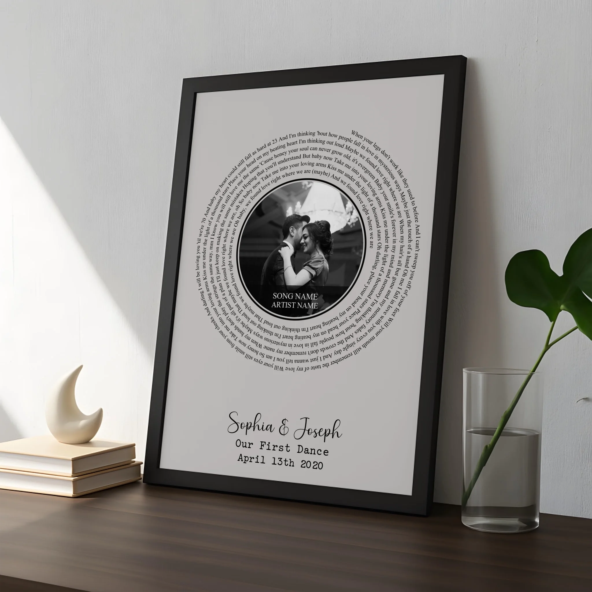 Black framed spiral lyrics print on dark wood surface next to a glass vase and moon ornament, personalised for Sophia and Jos