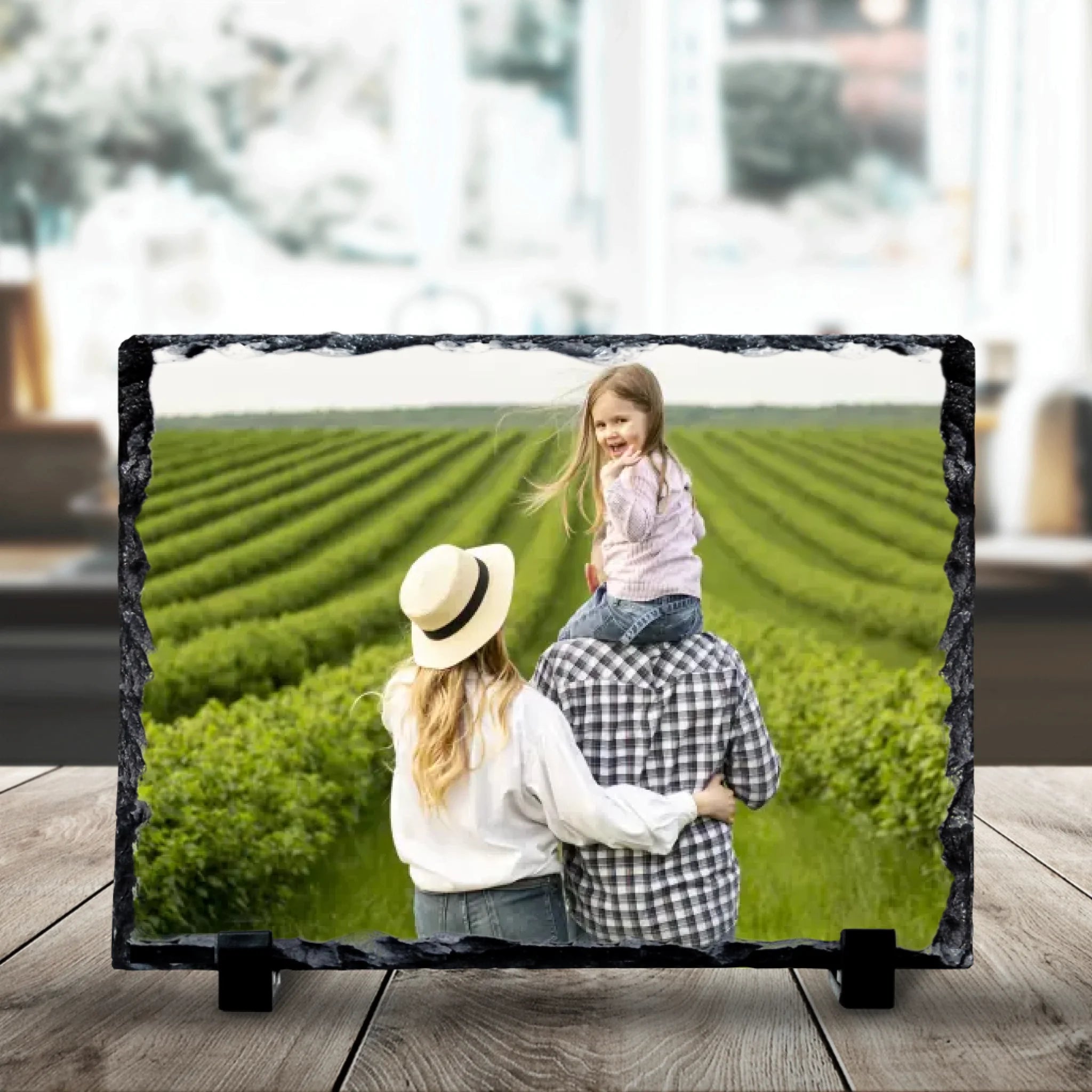 Personalised photo slate on a wooden surface showing a family of three facing green fields, with a young girl sitting on dad'