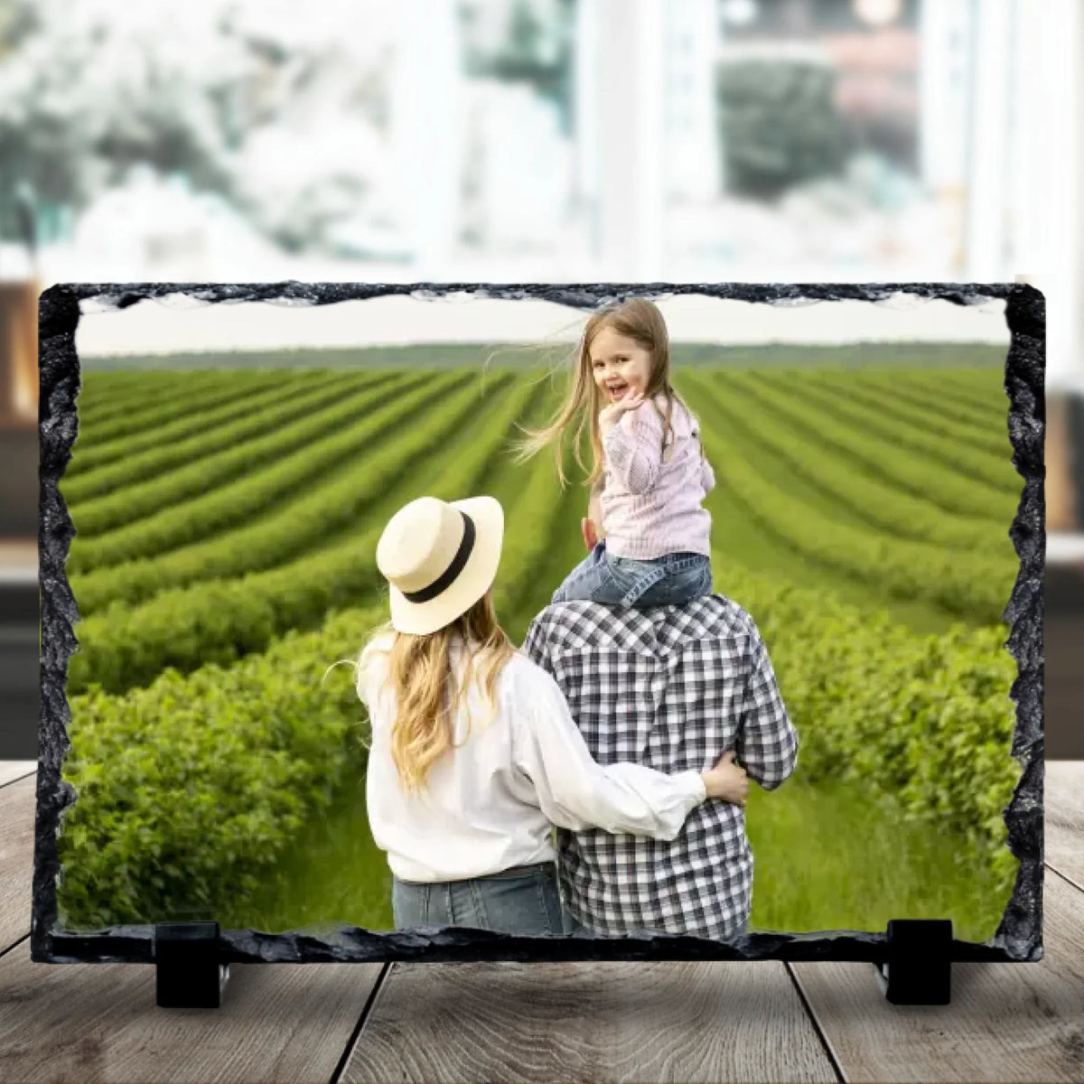 Close-up of a personalised photo slate displaying a family of three overlooking lush green rows of crops, daughter perched on