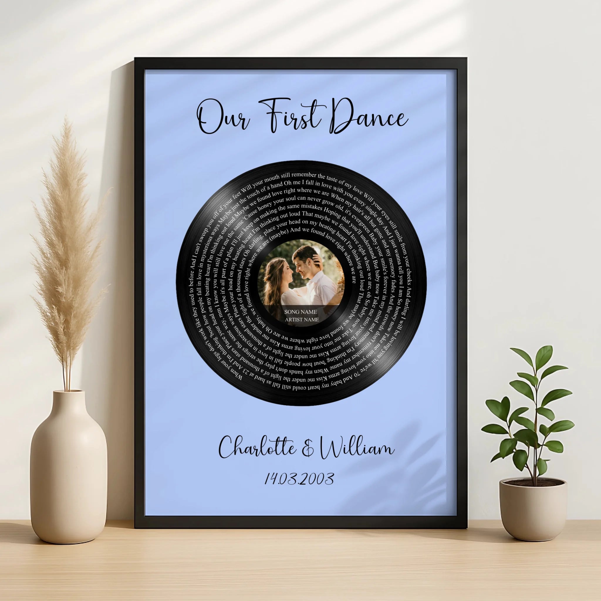 Black framed first dance vinyl record print on blue, couple photo at centre, displayed on wooden surface beside pampas grass 