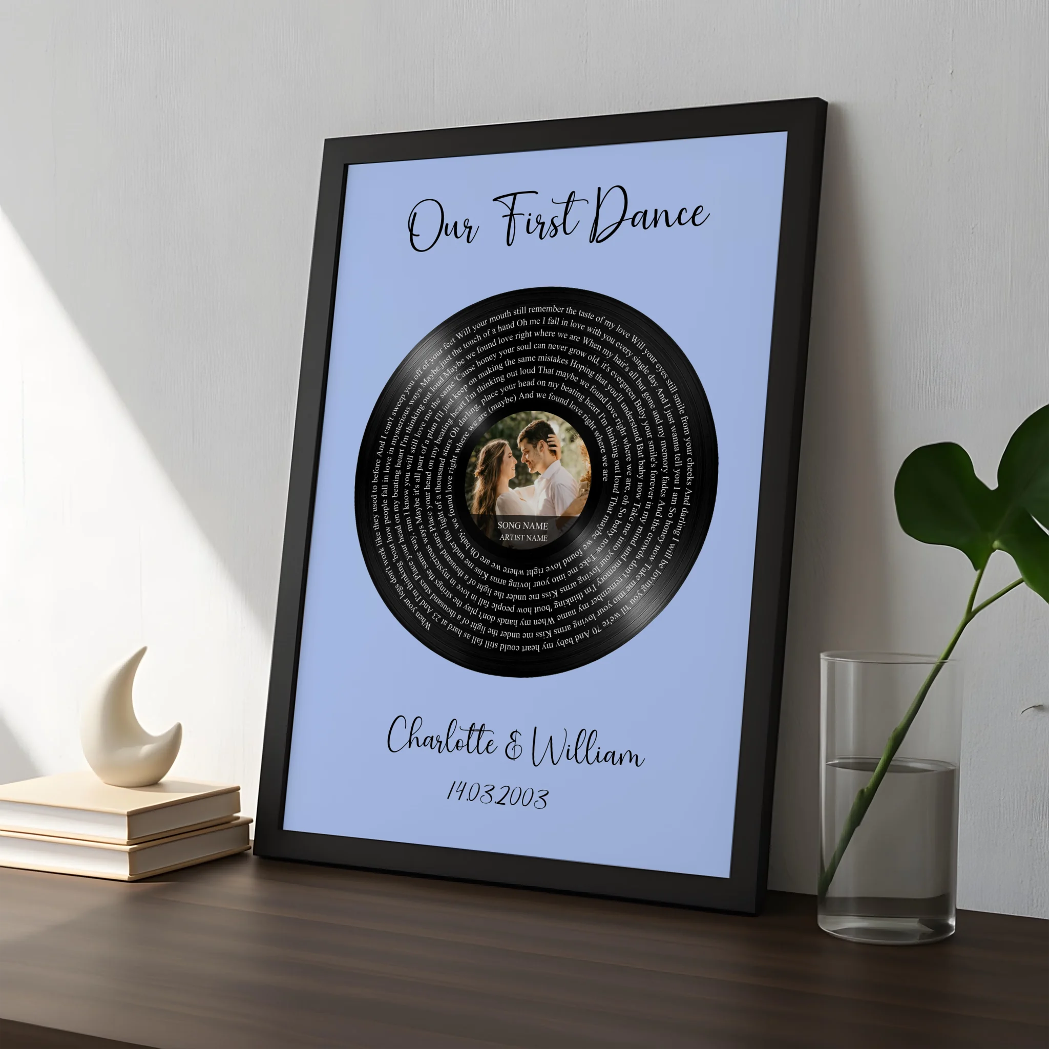 Black framed vinyl record song lyrics print on blue, leaning on dark wood surface beside a crescent ornament and glass vase w