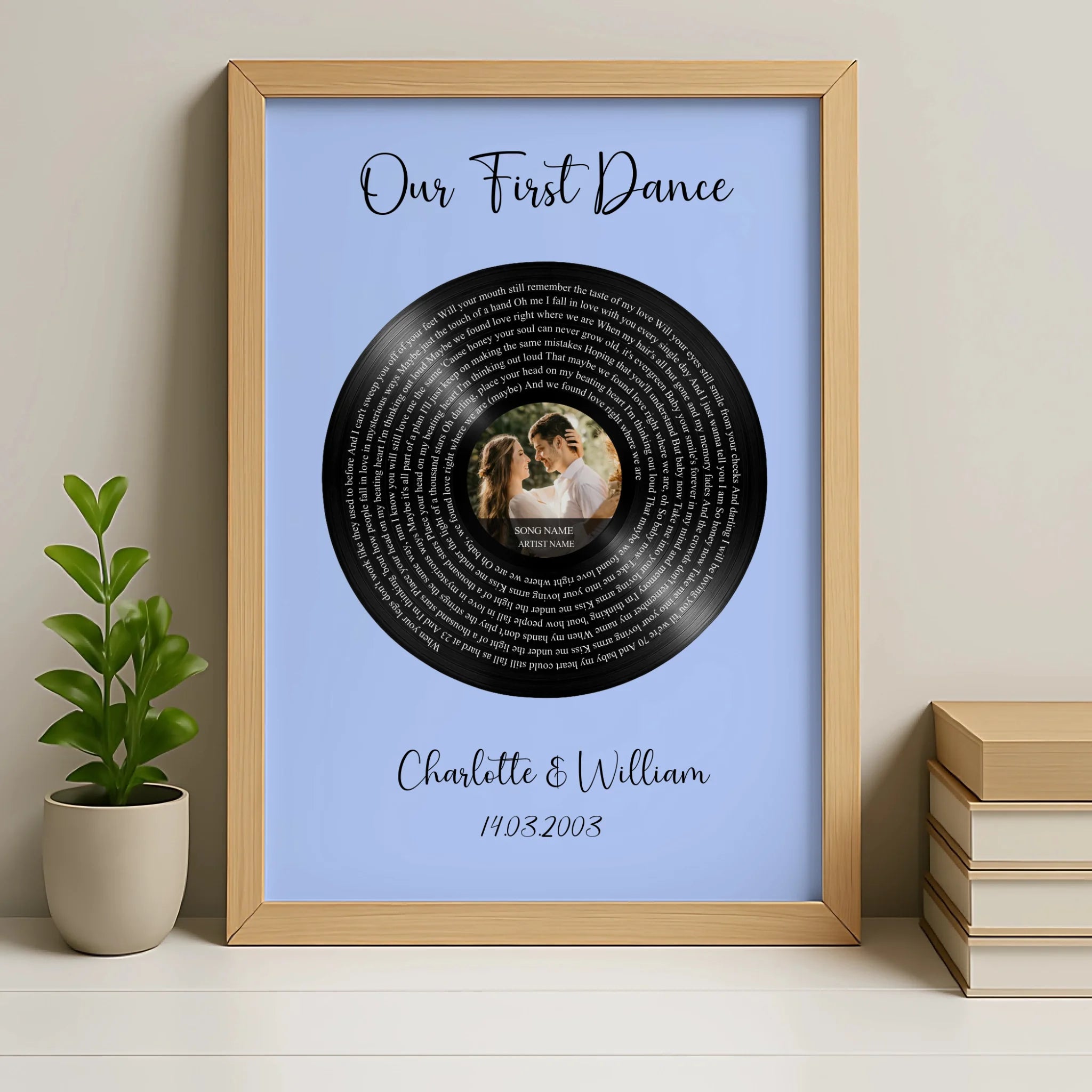 Natural wood framed first dance lyrics print on blue, vinyl record with couple's photo, beside a small potted plant and stack